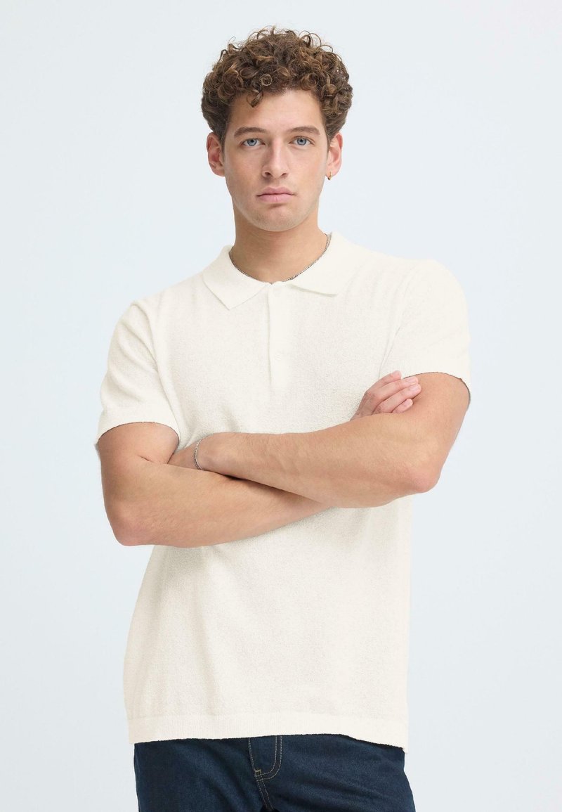 Νεαρός με σγουρά μαλλιά που φοράει λευκό πόλο μπλουζάκι με κοντά μανίκια και σκούρο τζιν, στέκεται με τα χέρια σταυρωμένα μπροστά σε λευκό φόντο.