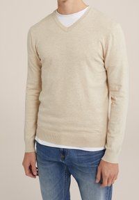 Hombre vistiendo un suéter V beige claro sobre una camisa blanca, combinado con jeans azules, de pie frente a un fondo neutral.