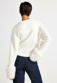 Kvinde set bagfra iført en hvid sweater med fuzzy manchetter og mørkeblå jeans mod en enkel lys baggrund.