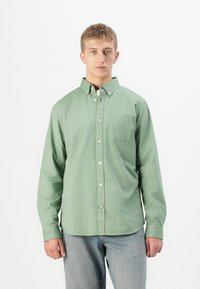 Un joven con el cabello corto está de pie vistiendo una camisa de botones de manga larga verde y jeans de color azul claro frente a un fondo blanco liso.