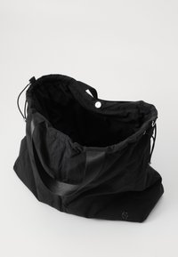 Schwarze Stofftasche mit gerafftem oberen Teil, ausgestattet mit zwei Griffen und einem Druckknopfverschluss. Das Innere ist geräumig und ungefüttert, mit einem dezenten Logo-Detail.