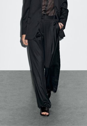 Person trägt lockere schwarze Hose, schwarzen Blazer, durchscheinendes dunkles Hemd, schwarze Sandalen mit offenen Zehen und ein silbernes Armband, geht auf einem grauen Boden.