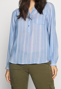 Blusa azzurro chiaro a maniche lunghe con strisce verticali bianche, chiusura a bottoni, colletto e una tasca sul petto. Tessuto leggero e strutturato.