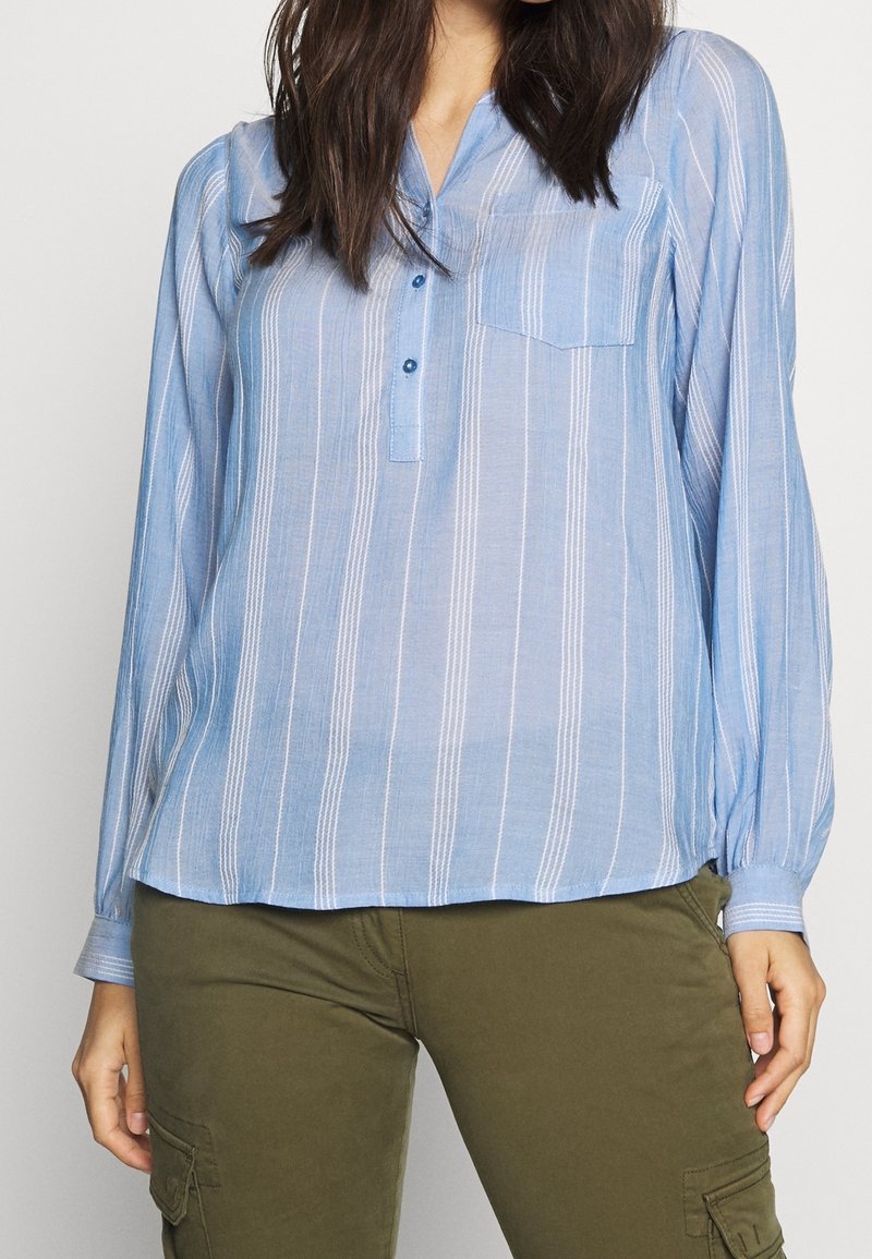 Blusa azzurro chiaro a maniche lunghe con strisce verticali bianche, chiusura a bottoni, colletto e una tasca sul petto. Tessuto leggero e strutturato.