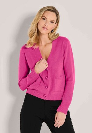 Frau mit langen blonden Haaren trägt eine hellpinkfarbene knöpfbare Strickjacke mit Taschen und schwarze Hosen und posiert vor einem hellen, einfarbigen Hintergrund.
