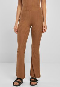 Des pantalons marron à côtes évasés présentent une taille haute, des lignes verticales pour la texture et des fentes à l'ourlet, associés à des sandales noires.
