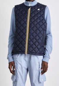 Marineblaue gesteppte Weste mit hohem Kragen, ausgestattet mit einem mehrfarbigen Reißverschluss und einem kleinen Logo. Getragen über einem hellblauen Pullover und einer Hose.