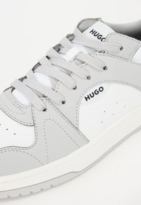Sneaker in pelle bianca con accenti grigi, punta tonda, lacci grigi e marchio "HUGO" sul lato. Suola testurizzata per una migliore aderenza.
