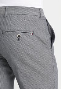 Graue gemusterte Hose aus strukturiertem Stoff, mit einer geknöpften Gesäßtasche und einem kleinen rot-weißen-blauen Akzent in der Nähe der Tasche.