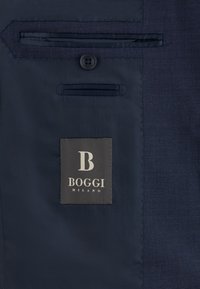 Intérieur de costume bleu marine avec poche boutonnée, étiquette cousue portant la mention "B Boggi Milano" et tissu texturé sur le côté droit.