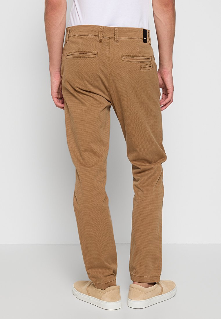 Slim-Fit, braune Baumwollhose mit subtiler Textur, zwei Gesäßtaschen und einem schwarzen Etikettakzent an der Taille, kombiniert mit beigen Slippern.