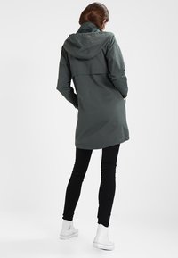 Femme portant un manteau gris à capuche, un pantalon noir et des baskets blanches montantes, se tenant avec les mains dans les poches, tournée vers le dos.