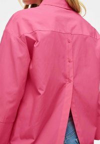Camicia rosa realizzata in tessuto morbido, con chiusura a bottone sul retro e design a spacco per un tocco extra. Collo strutturato e vestibilità comoda.
