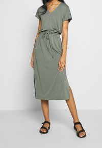 Mujer vistiendo un vestido midi verde oliva con cintura ajustable y mangas cortas, combinado con sandalias de tiras negras sobre un fondo gris.