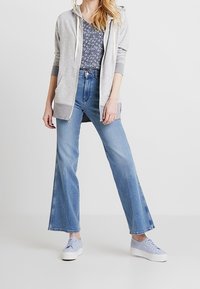 Felpa grigia chiara con zip e tasche frontali, indossata sopra un top navy a fantasia. Jeans blu a gamba larga e sneakers platform azzurre completano l'outfit.