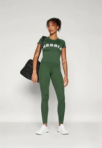 Zeleni set sportske odjeće sastoji se od kratkotrajne majice s natpisom "NEBBIA" u bijeloj boji i usklađenih tajica. Uključuje crnu torbu za teretanu i bijele tenisice.