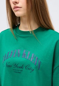 Νέος με ίσια καστανά μαλλιά φορώντας ένα πράσινο φούτερ Calvin Klein με την επιγραφή «Νέα Υόρκη».