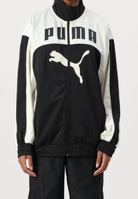 Schwarze und weiße Zip-Jacke mit hohem Kragen, die das "PUMA"-Logo und eine springende Katzen-Grafik zeigt. Glattes Material mit elastischen Bündchen und Saum.