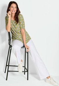 Groen patroon top met korte mouwen, gecombineerd met witte wijde broek en witte sneakers. Model zit op een zwarte metalen kruk.