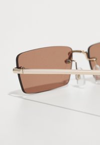Lunettes de soleil rectangulaires teintées marron avec une fine monture en métal doré et des accents noirs, dotées de coussinets de nez transparents et de quincaillerie vissée.
