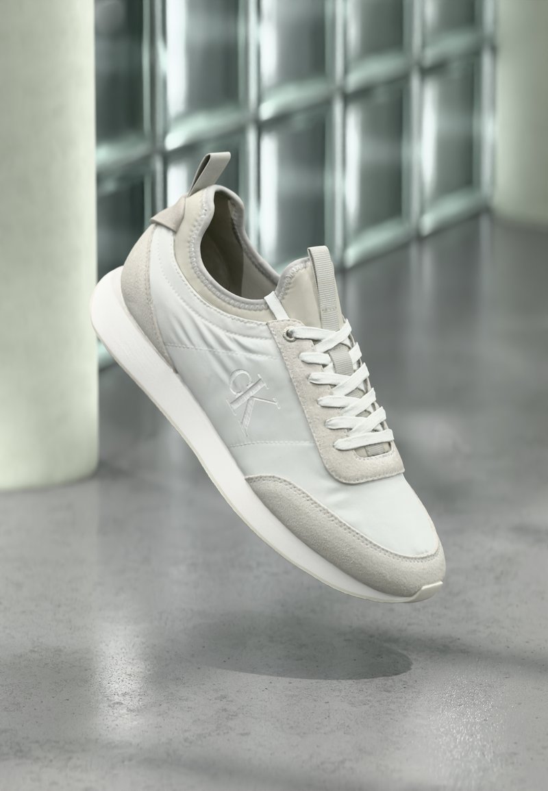 Λευκό και μπεζ αθλητικό παπούτσι με κορδόνια και λογότυπο "ck" στο πλάι, εμφανιζόμενο στον αέρα με φόντο ένα θολωμένο σύγχρονο υπόβαθρο από γυαλί και σκυρόδεμα.