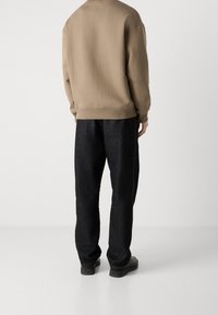 Uomo con felpa beige, pantaloni neri larghi e scarpe nere, in piedi con le spalle rivolte verso la macchina fotografica su uno sfondo bianco semplice.