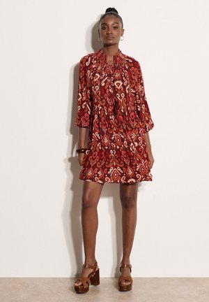 Kobieta stojąca w czerwonej, wzorzystej sukience do kolan z rękawami do połowy przedramienia i brązowych platformach, na jednolitym jasnym tle.