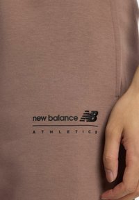 Camiseta deportiva marrón con un logotipo bordado en negro de "New Balance" y el texto "ATHLETICS", con una textura de tela suave y un diseño ajustado.