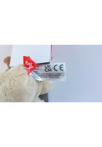 Beige plush teddy bear with soft fur, featuring a red label and a white tag displaying care instructions and safety certifications.