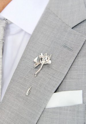 Srebrna przypinka w kształcie kwiatu z polerowanymi płatkami i szczegółowo wykonanym łodygą, przypięta do jasnoszarego materiału garnituru z białą poszetką.