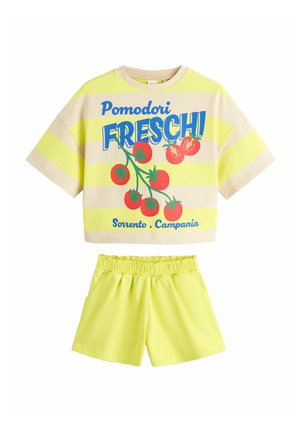 Krótki rękaw w żółto-beżowe paski z grafiką czerwonego pomidora i niebieskim napisem, zestawiony z jednolitymi żółtymi szortami z elastycznym pasem.