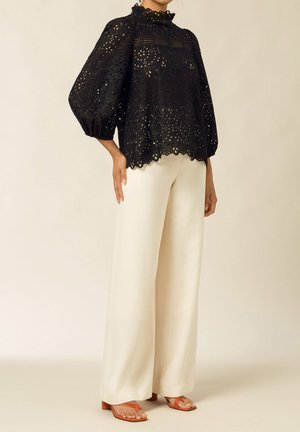 Blouse noire à œillets avec un col haut à volants et des manches ballons amples, associée à un pantalon large crème et des sandales orange.