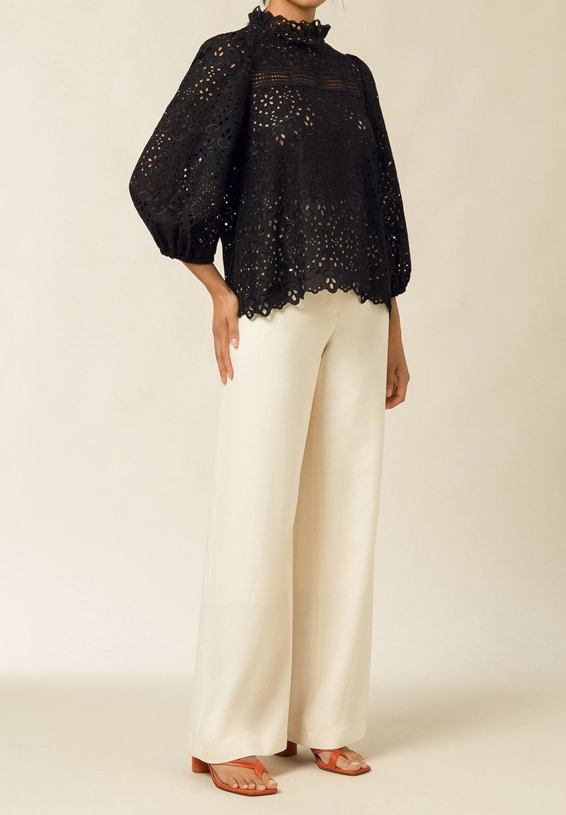 Blusa nera con occhielli, collo alto arricciato e maniche a palloncino morbide, abbinata a pantaloni larghi beige e sandali arancioni.