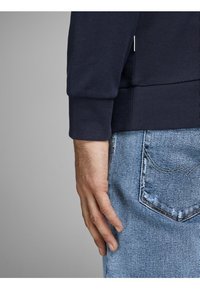 Felpa blu navy con polsini a costine, abbinata a jeans in denim azzurro chiaro con dettagli di cucitura sulle tasche posteriori. Mano posizionata vicino alla vita.