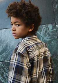 Ragazzo con capelli ricci che guarda indietro oltre la spalla, indossando una camicia a quadri con "Brooklyn" ricamato sulla schiena, in piedi accanto a un muro di pietra blu.