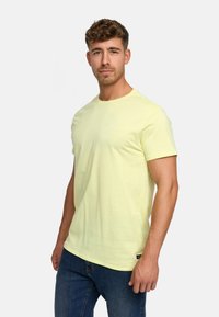 Helles gelbes Baumwoll-T-Shirt mit Rundhalsausschnitt und kurzen Ärmeln. Verfügt über ein kleines Logo auf der Brust und ein Etikett am Saum, kombiniert mit blauen Jeans.