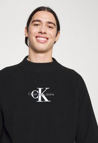 Schwarzer gerippter Pullover mit hohem Kragen und gesticktem weißen Logo "Calvin Klein Jeans" unter einem auffälligen "CK" in moderner Schriftart.
