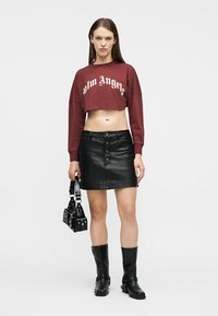 Γυναίκα με κοντό μπορντό φούτερ, μαύρη δερμάτινη φούστα με κουμπιά μπροστά, μαύρες μπότες και κρατώντας μια μαύρη τσάντα με διακοσμητικά καρφιά.