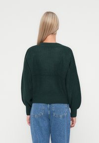 Ciemnozielony sweter dzianinowy z okrągłym dekoltem, obszernymi rękawami i ściągaczem, zestawiony z dżinsami w średnim odcieniu o wysokim stanie.