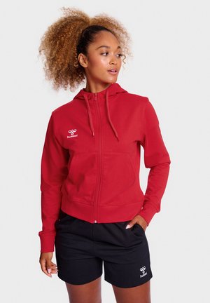 Rode zip-up hoodie met een capuchon, lange mouwen en kangoeroe zakken. Voorzien van een wit logo op de borst. Gekleed met zwarte shorts.