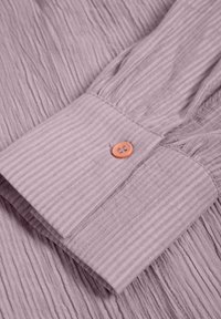 Manche en tissu texturé violet clair avec des plis verticaux. Présente un bouton orange contrastant au poignet.