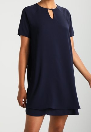Donna che indossa un vestito blu navy a maniche corte con un piccolo taglio a goccia sul collo e un orlo a strati.