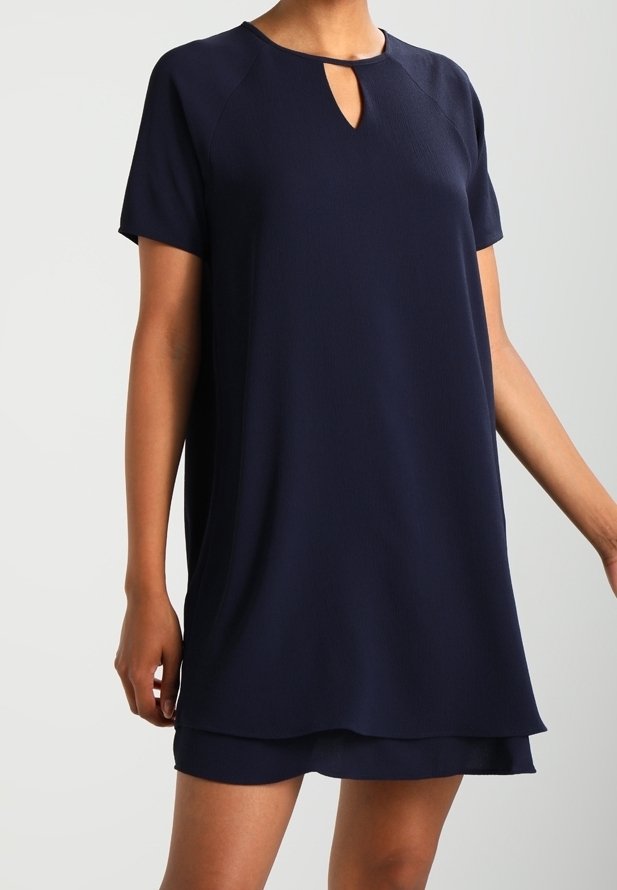 Femme portant une robe bleu marine à manches courtes avec une petite découpe en forme de goutte d'eau au niveau de l'encolure et un ourlet à volants.