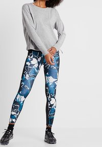 Grå långärmad tröja med logga på ärmen, kombinerad med blå och vita mönstrade leggings. Leggingsen har svarta sidpaneler och ankeldetaljer.