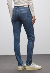 Osoba ubrana w niebieskie dżinsy o białych paskach po bokach oraz białe trampki, stojąca na szarym podłożu w tle białej ściany.