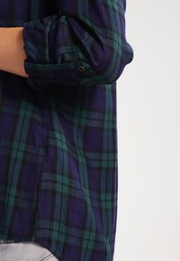 Chemise en flanelle avec un motif tartan violet et vert. Elle présente des manches retroussées et un détail de bouton au niveau du poignet. Tissu doux et texturé.