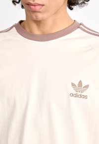 Κρέμα βαμβακερό μπλουζάκι με καφέ ριγωτό γιακά και τρεις ρίγες στους ώμους. Διαθέτει κεντημένο λογότυπο Adidas στο στήθος.