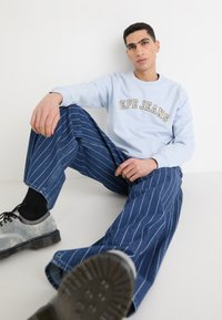 Sudadera azul claro con el logo de "Pepe Jeans", combinada con jeans de pierna ancha a rayas azul. Zapatos grises con suelas negras y calcetines oscuros.
