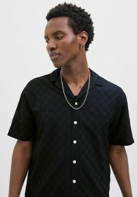 Chemise noire à manches courtes avec un motif à carreaux texturé, dotée d'un col et de boutons blancs, complétée par un collier en chaîne argenté.