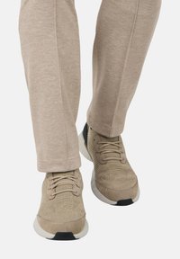 Šviesiai rudos spalvos kelnių su baltais batu, pagamintais iš megztinio, vaizdas nuo šoninės pusės iki vidurinės blauzdos baltoje fone.